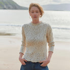 A woman standing on a sandy beach wearing a textured cream-colored knit sweater and blue jeans, with the ocean and distant hills in the background.