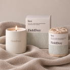 A FieldDay 'Sea' scented vegetable wax candle set, featuring a lit cream-colored glass jar, its kraft paper wrapped wax refill container, and the product box, all styled on a textured beige linen fabric against a neutral beige background.
